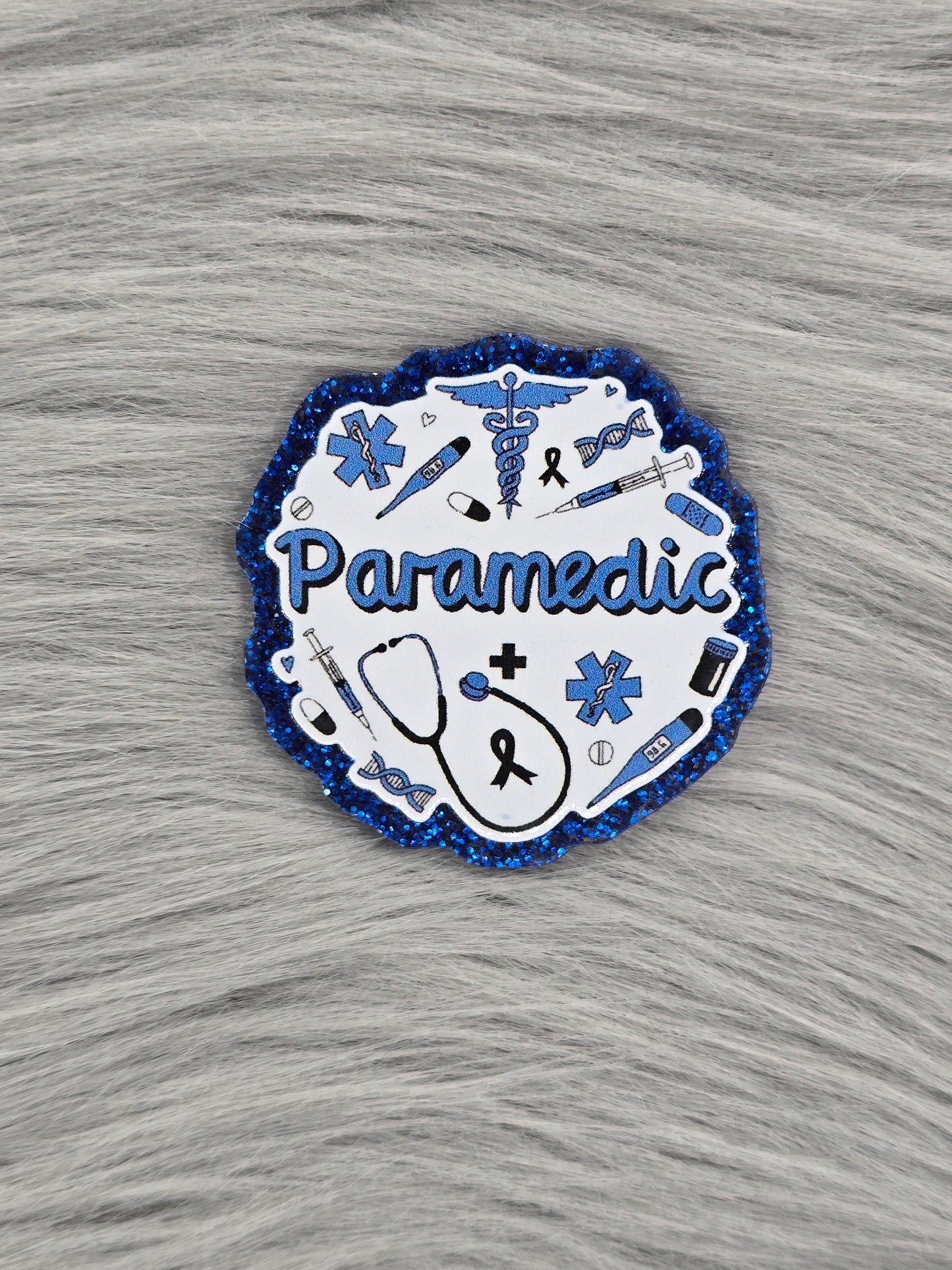 Glitter blue background acrylic flatback with white picture that says 'Paramedic' in blue with medical supplies in blue and black