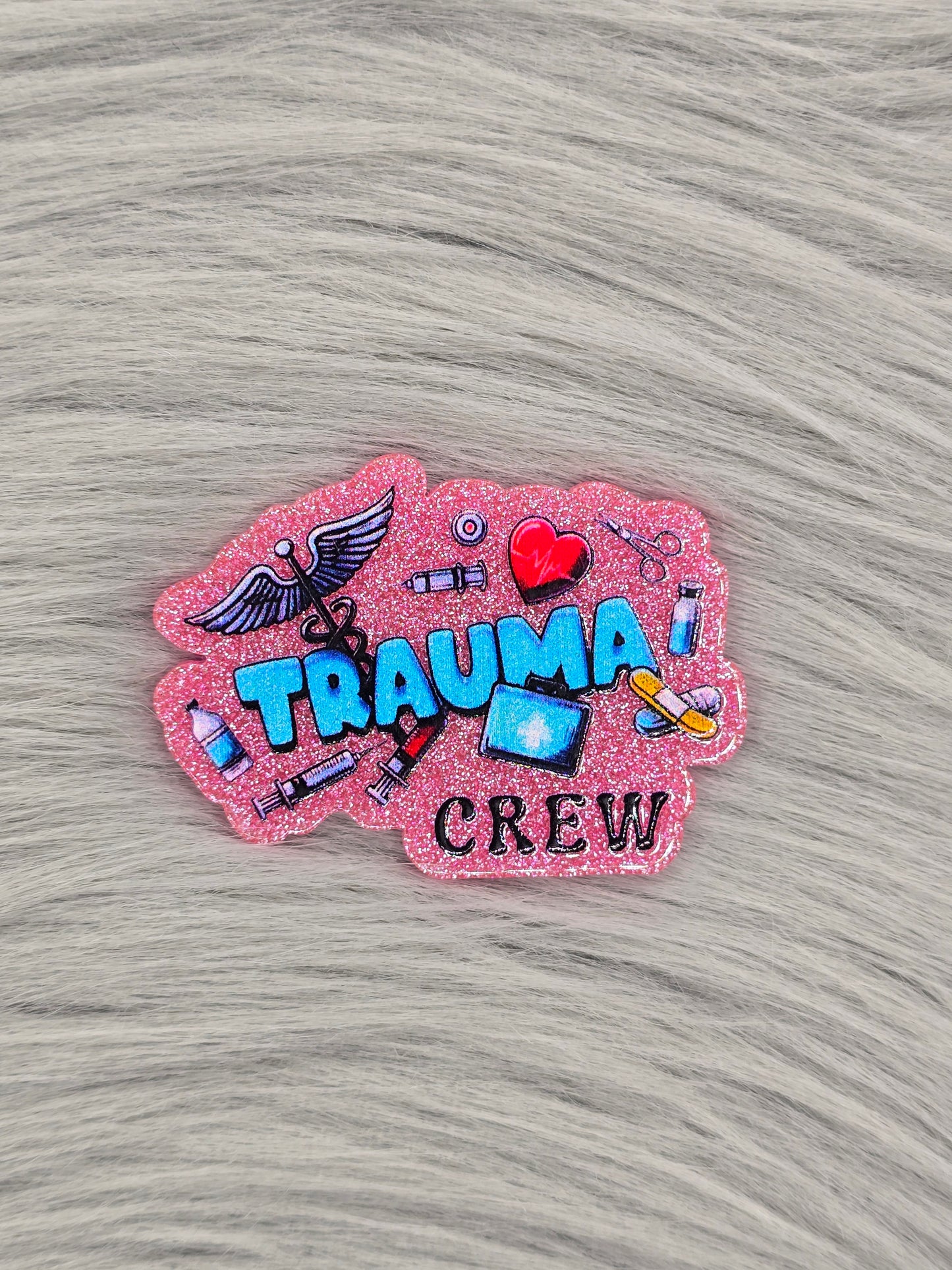 Pink glitter acrylic flatback with the words in blue hat say "trauma crew" with medical pictures