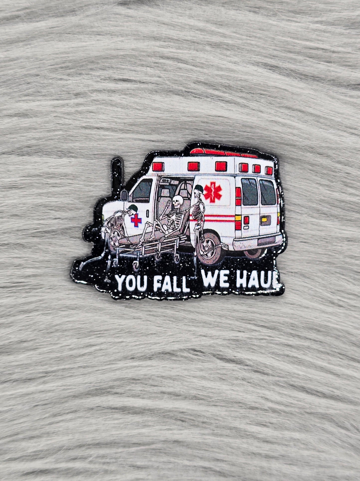 Black glitter acrylic flatback with an ambulance and 3 skeletons, at the bottom in white it says 'you fall we haul'