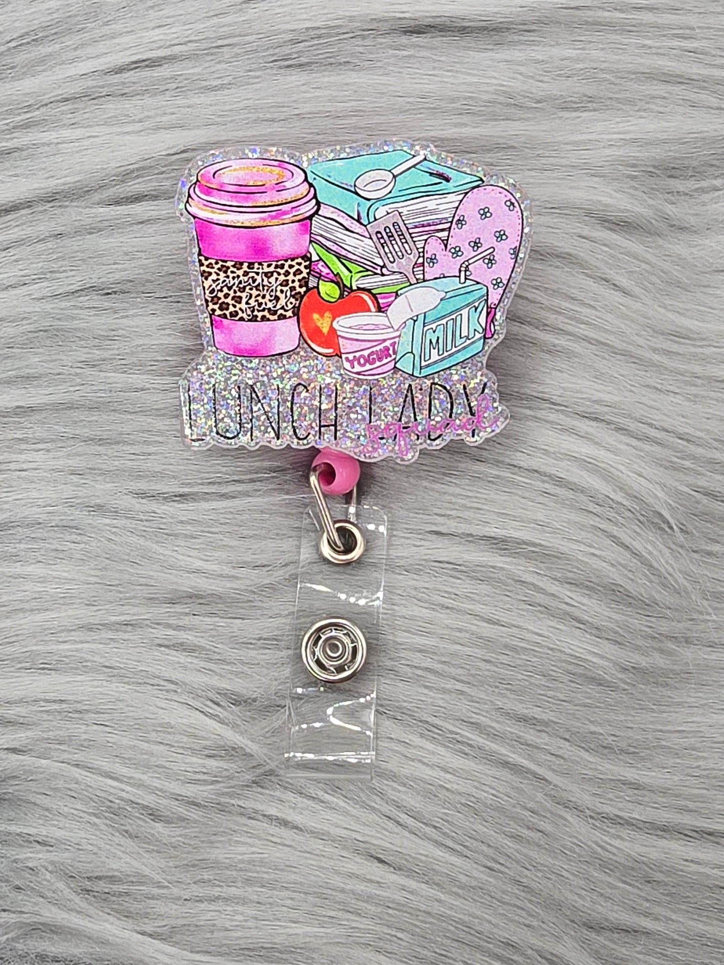 Silver background of an acrylic flatback that says Lunch Lady Squad with lunch items on a pink badge reel