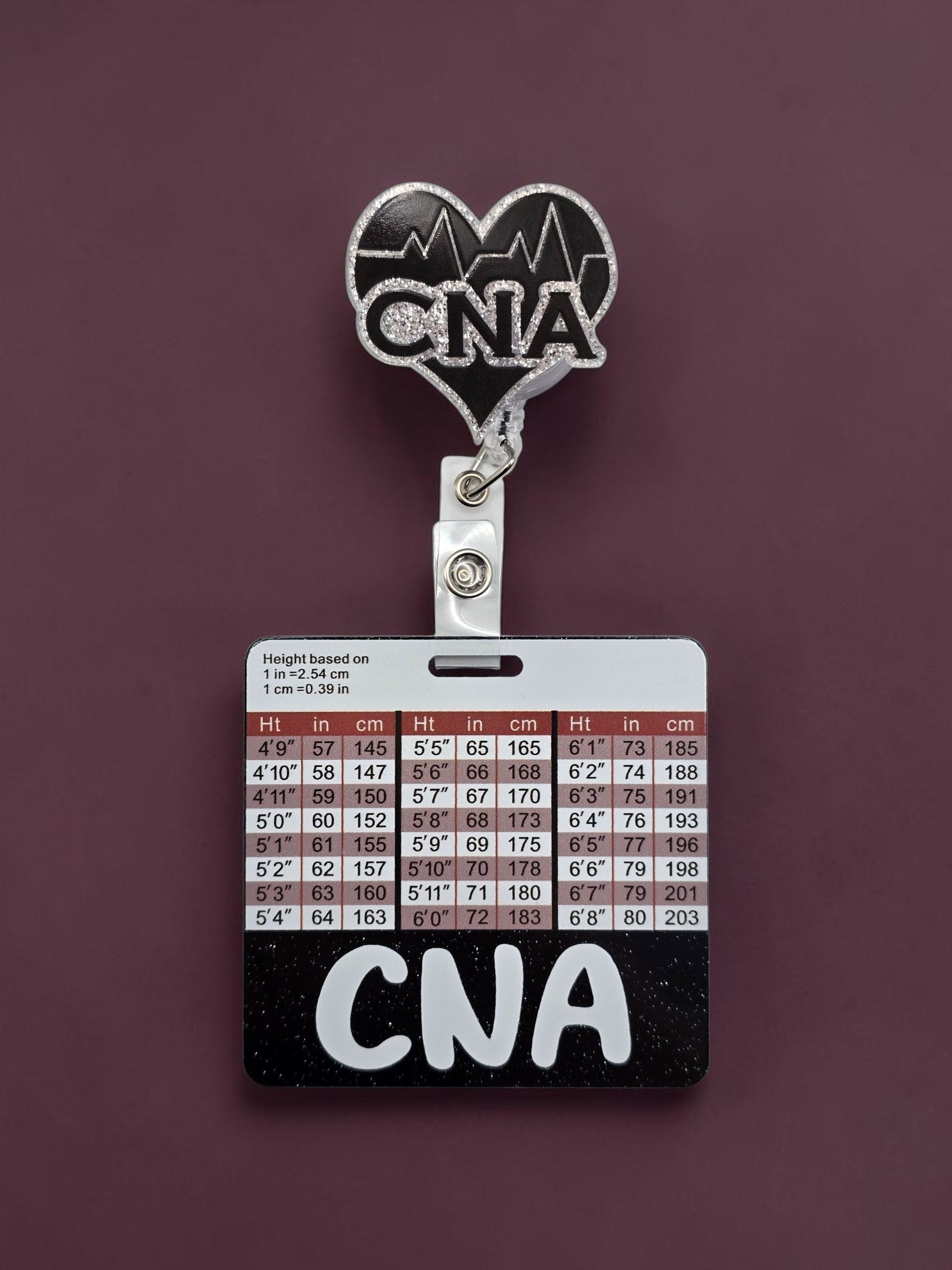A flatback that is heart shape with the letters CNA in the middle attached to a badge reel and a square badge buddy with height and weight and black strip at the bottom with the letters CNA in white in the middle