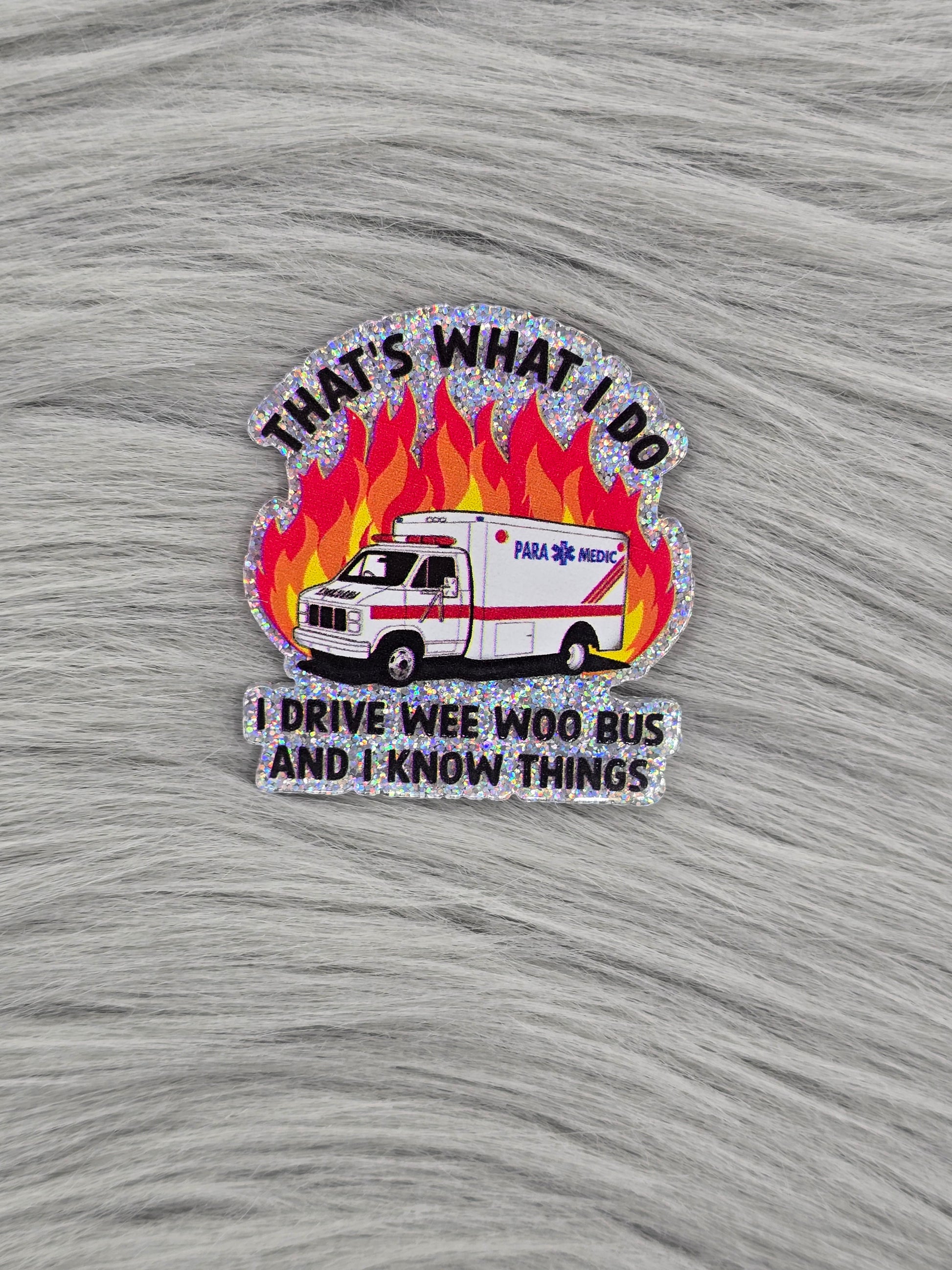 Silver glitter background acrylic flatback with a picture of an ambulance with flames behind it and the words "That's What I Do I Drive The Wee Woo Bus And I Know Things"