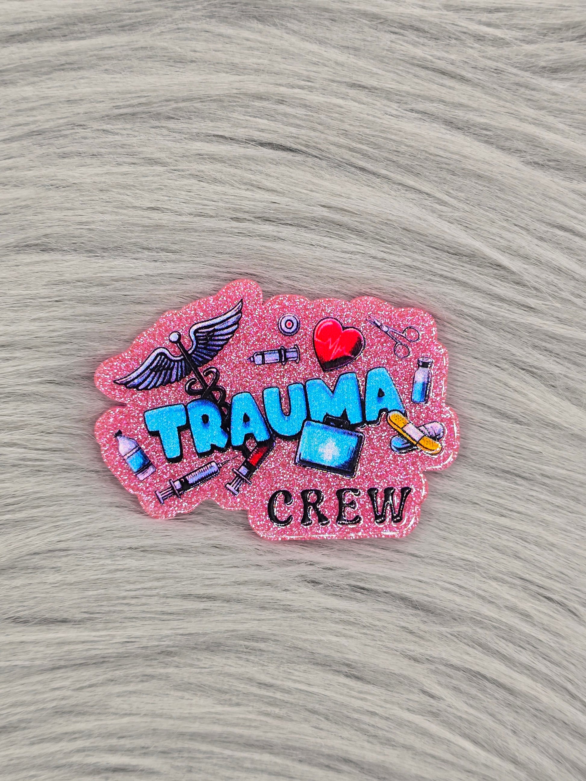 Pink glitter acrylic flatback with the words in blue hat say "trauma crew" with medical pictures