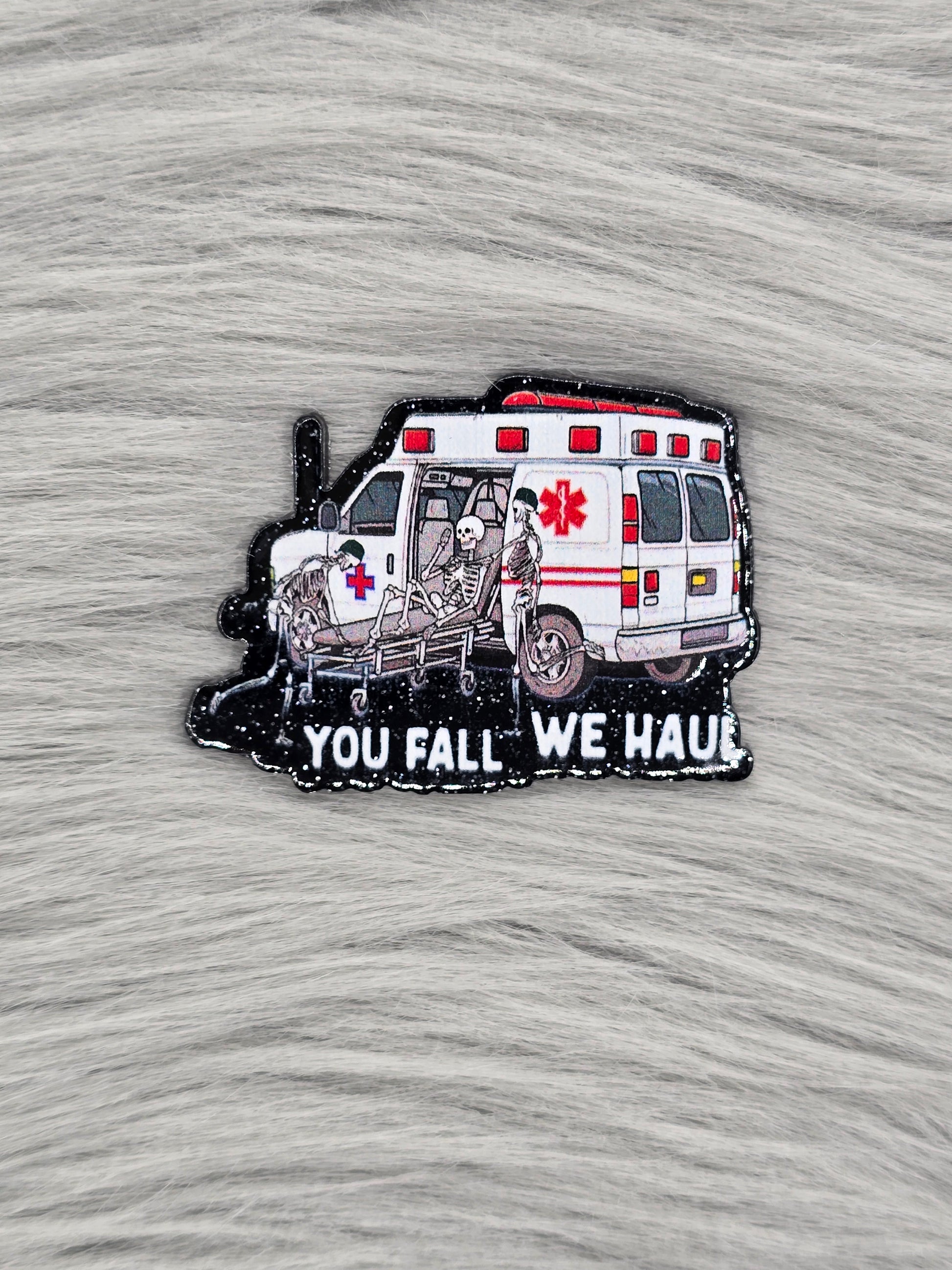 Black glitter acrylic flatback with an ambulance and 3 skeletons, at the bottom in white it says 'you fall we haul'