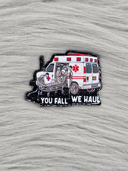 Black glitter acrylic flatback with an ambulance and 3 skeletons, at the bottom in white it says 'you fall we haul'