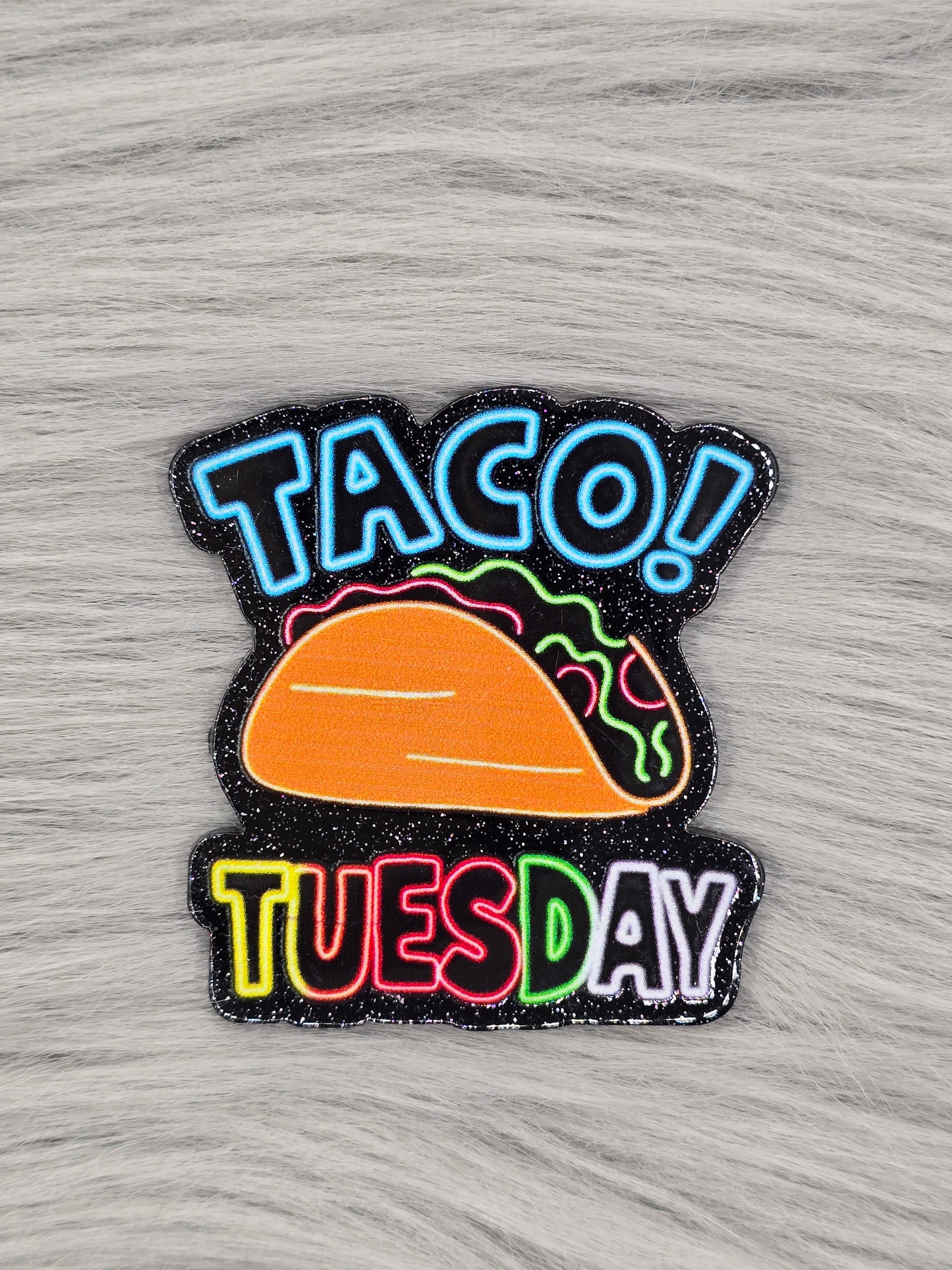 Black glitter flatback with the word "TACO!" in light blue at the top and a taco in the middle and the word "Tuesday" at the bottom in rainbow colors