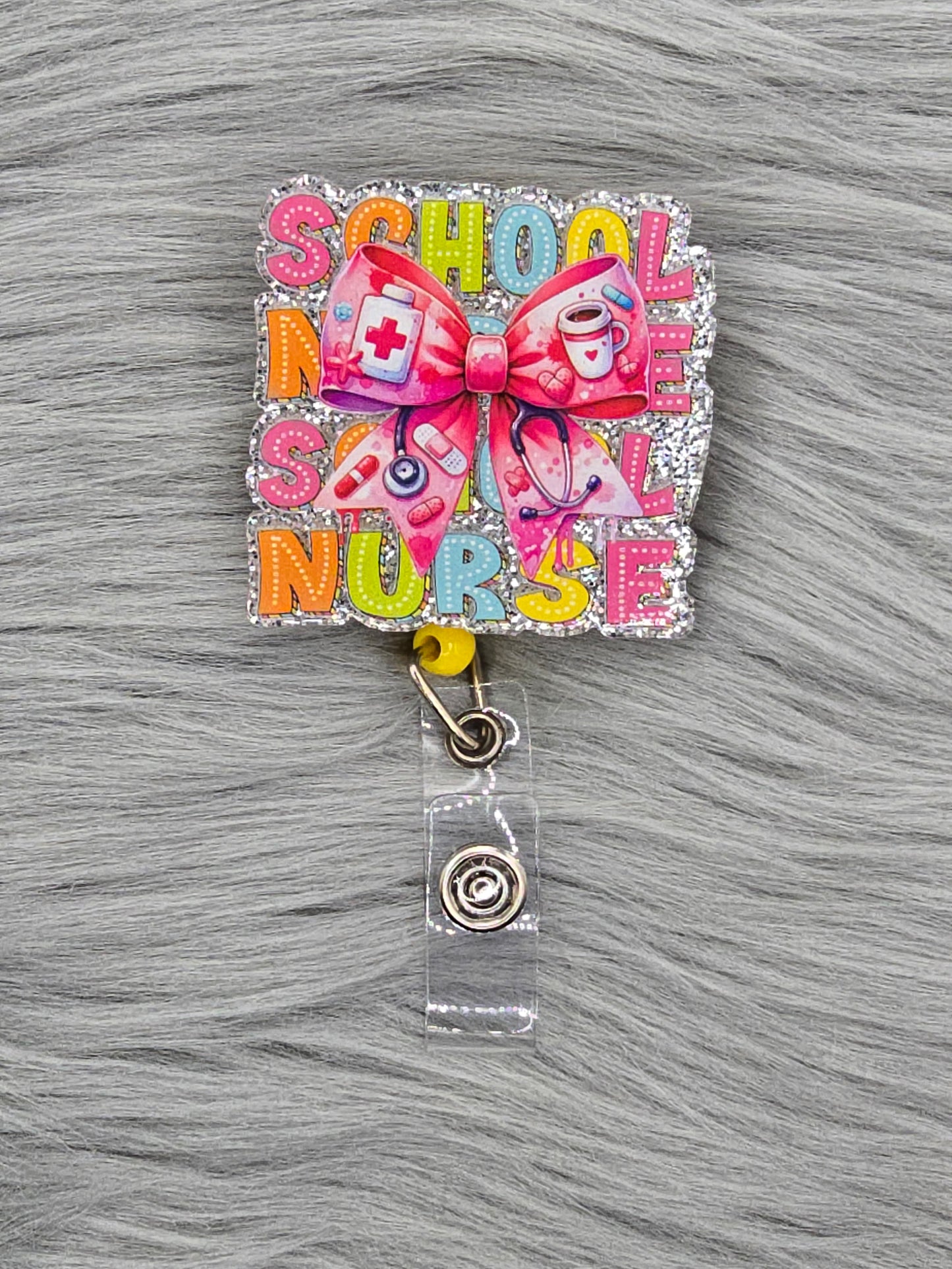 Acrylic flatback that says school nurse in colorful colors with a pink medical bow in the middle on a yellow badge reel