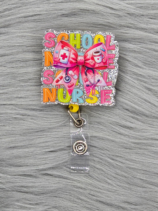 Acrylic flatback that says school nurse in colorful colors with a pink medical bow in the middle on a yellow badge reel