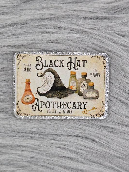acrylic flatback with a light yellow background with black words that say " Black Hat Apothecary" It has a witches had and potion bottles