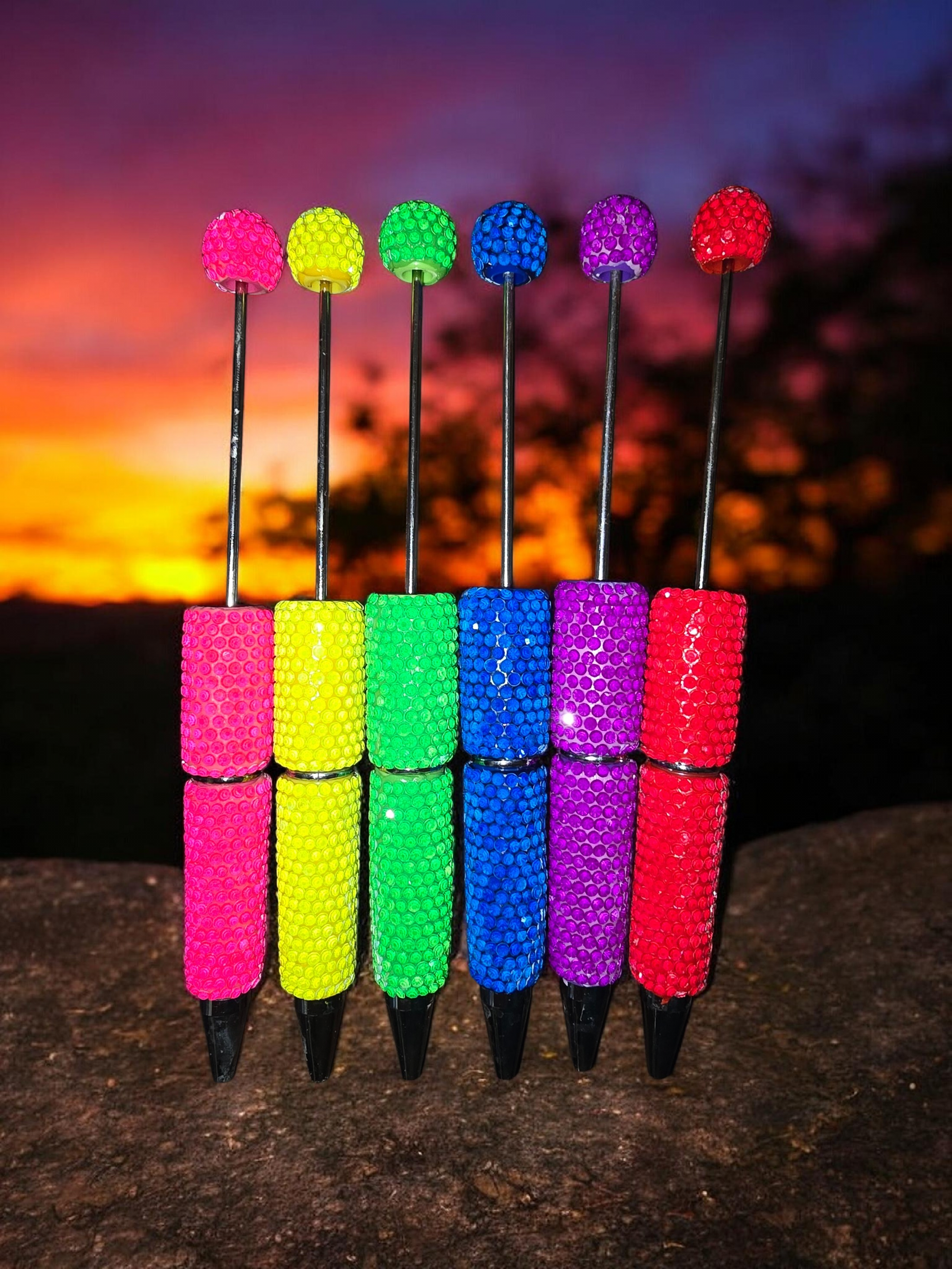 Colorful beadable pens in neon pink, yellow, green, blue, purple and red against a sunset background