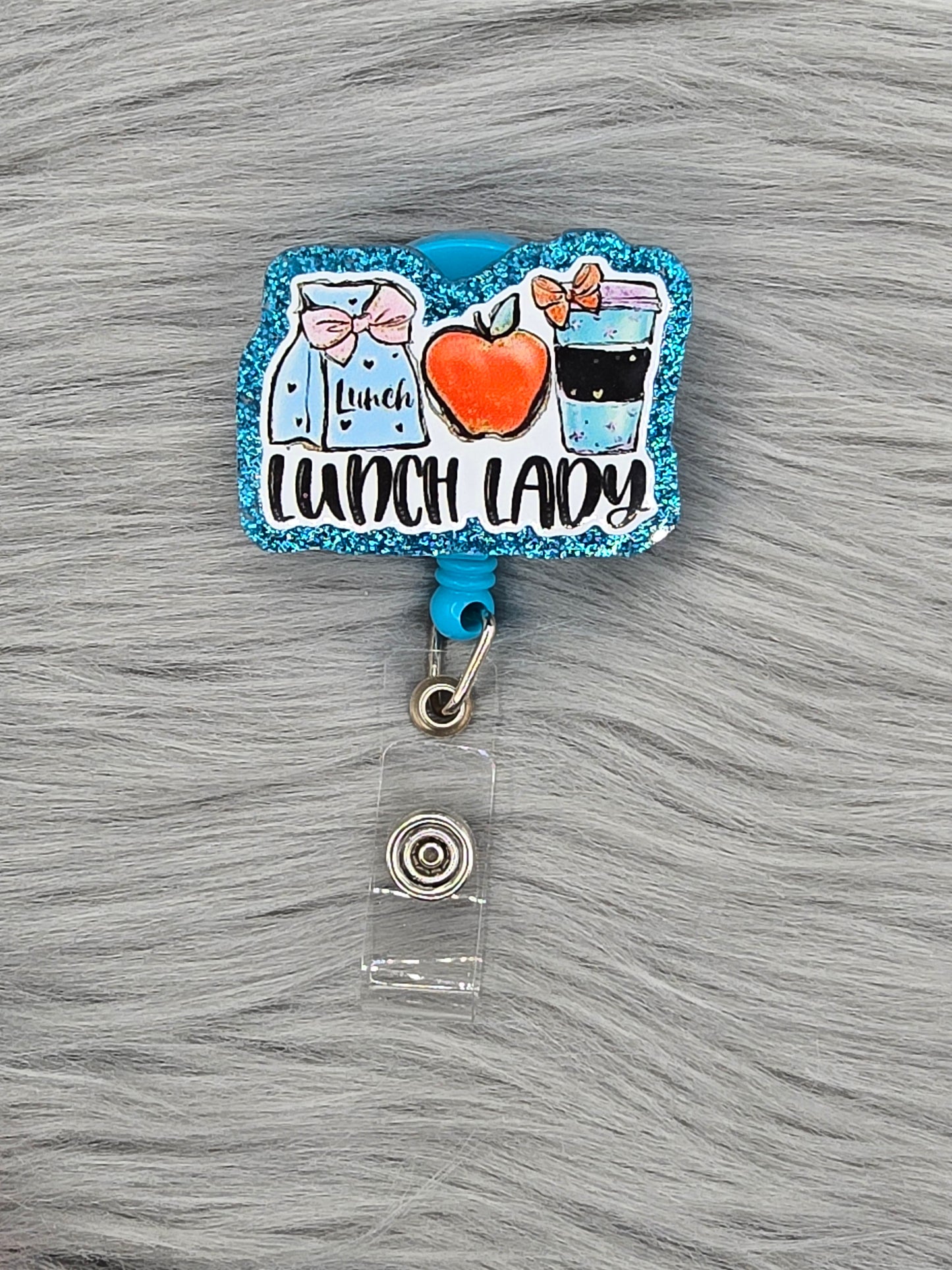 Blue acrylic flatback with the words Lunch Lady and pictures of an apple, sack lunch and coffee cup on a light blue badge reel