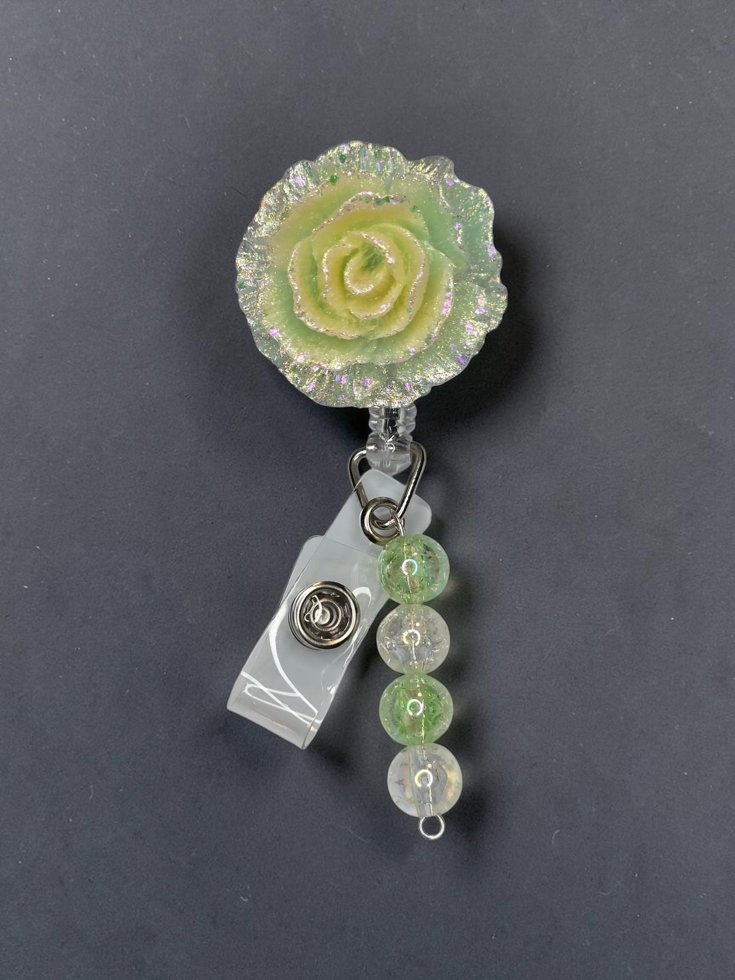 A close-up of a retractable badge reel with a textured light green and yellow rose charm, a clear plastic clip, and three hanging green and clear faceted beads on a dark gray background.