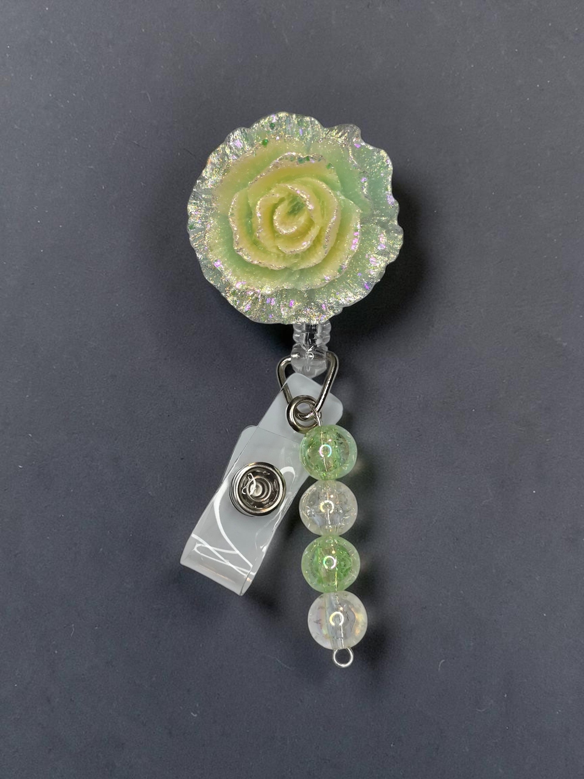 A close-up of a retractable badge reel with a textured light green and yellow rose charm, a clear plastic clip, and three hanging green and clear faceted beads on a dark gray background.